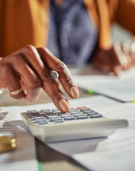 Close up of african american woman hands calculating tax while working at desk. Casual businesswoman using calculator to check monthly income and expenses of her freelance activity. Mature lady calculating business balance and prepare tax reduction with stacks of coins on desk.