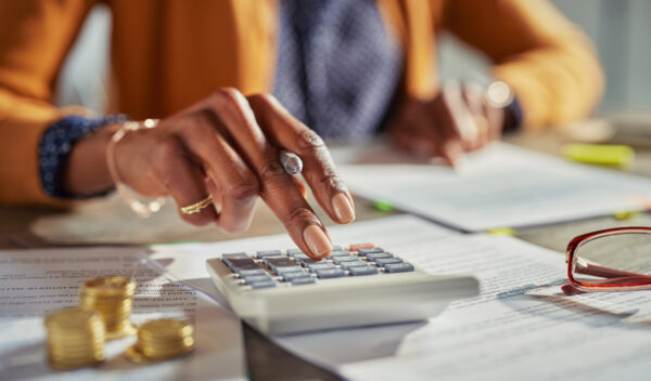 Close up of african american woman hands calculating tax while working at desk. Casual businesswoman using calculator to check monthly income and expenses of her freelance activity. Mature lady calculating business balance and prepare tax reduction with stacks of coins on desk.