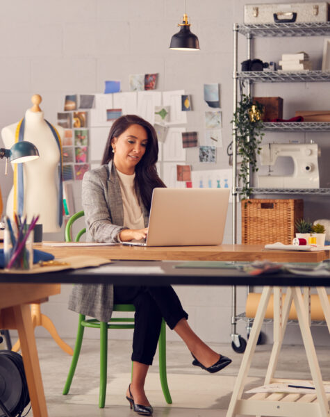 Female Fashion Designer In Studio Working On Sketches Or Documents At Desk With Laptop