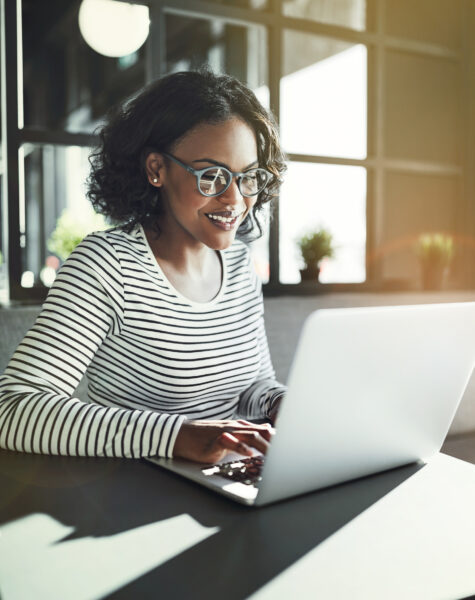 Smiling young African woman wearing glasses browsing online with a laptop while sitting alone at a table